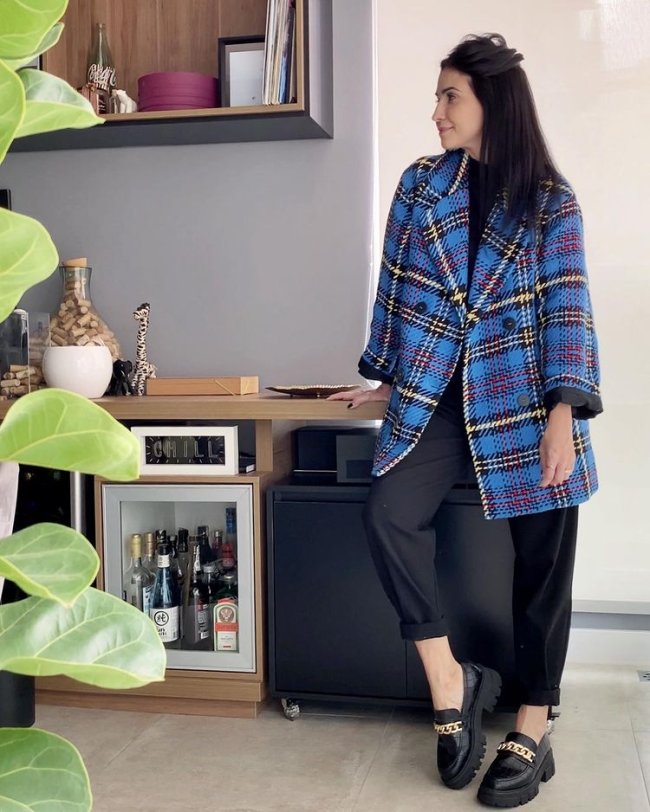 Looks com mocassim Jovem posando em sua cozinha, olhando para o lado, usando sobretudo xadrez azul com calça preta e mocassim.