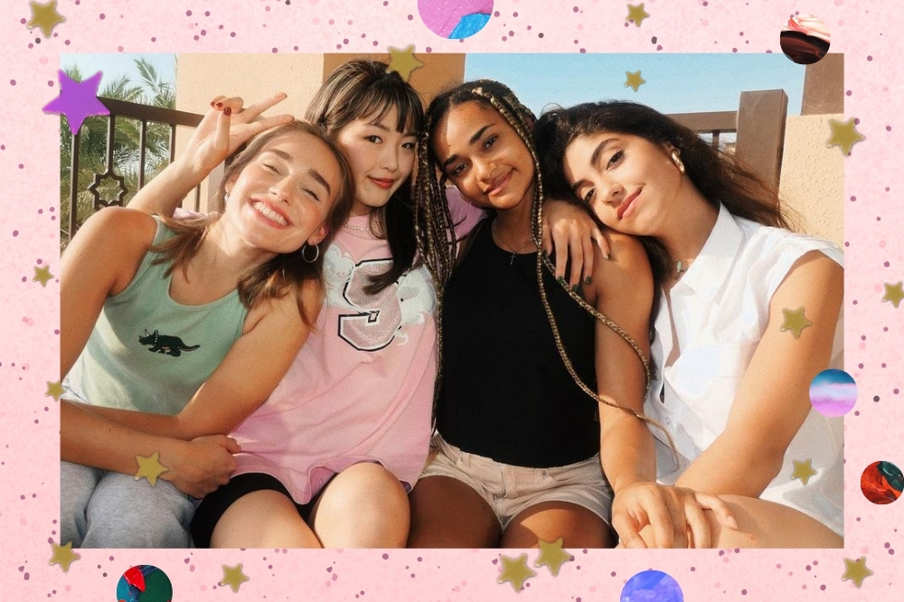 Foto com as integrantes do grupo do Now United, Sina, Hina, Mélanie e Nour sentadas uma ao lado da outra e abraçadas, todas sorridentes e Hina faz o sinal de paz e amor com uma das mãos.