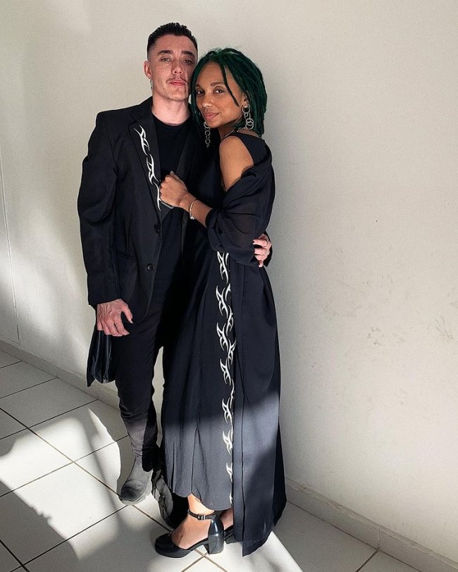 Nataly Neri e namorado Casal encostado em parede branca abraçado usando roupas pretas. Os dois tem uma expressão séria.