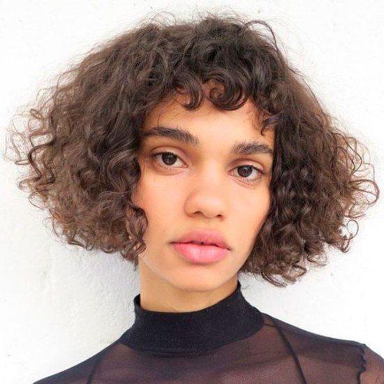 baby bangs cabelo cacheado Foto de uma mulher. Ela usa blusa preta de gola alta, cabelo curto e cacheado com baby bangs e pele natural. Ela olha para a câmera e não sorri.