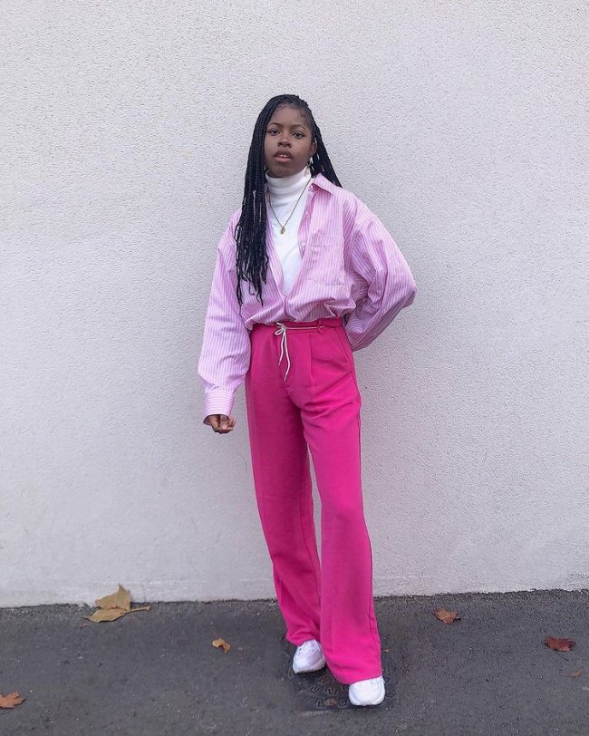 Foto de uma mulher na rua. Ela usa uma blusa de gola alta branca, camisa rosa com listras brancas aberta, calça rosa e tênis branco. Ela olha para a câmera e não sorri.