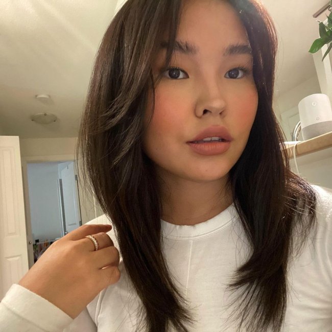cabelo curtain bangs Selfie de uma mulher. Ela usa uma blusa de manga longa branca, cabelo solto com franja cortininha. Ela está com a mão direita apoiada no ombro direito, olha para a câmera e não sorri.