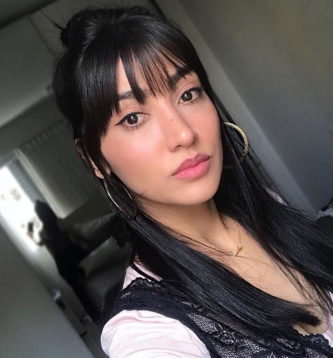 cabelo franja desfiada Selfie de uma mulher. Ela usa uma blusa cor creme com detalhes de renda preta, maquiagem natural, brinco de argola e cabelo com franja desfiada solto. Ela olha para a câmera e não sorri.
