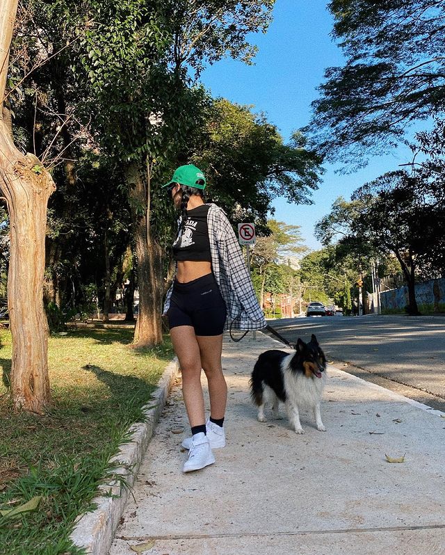 camisa xadrez com shorts Foto de uma mulher em uma calçada com um cachorro. Ela usa uma camiseta preta, shorts preto, camisa xadrez, boné verde e tênis branco. Ela olha para trás.