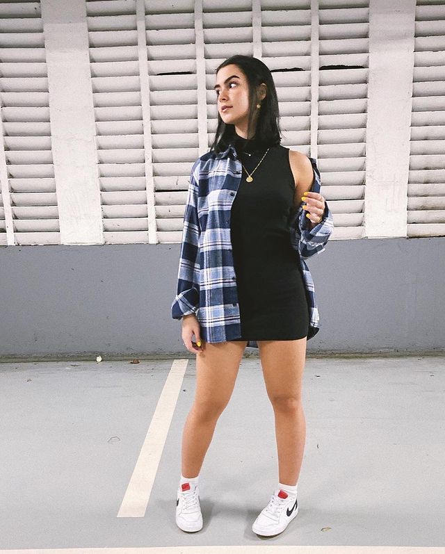 camisa xadrez com vestido preto Foto de uma menina em um estacionamento. Ela usa um vestido preto de gola alta e uma camisa xadrez oversized como sobreposição. Ela olha para o lado e não sorri.