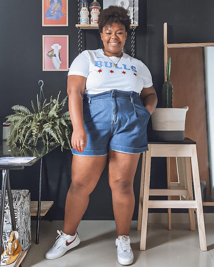 Garota usando camiseta branca, short jeans de cintura alta e tênis branco com detalhe em vermelho. Ela está sorrindo e com uma das mãos no bolso.