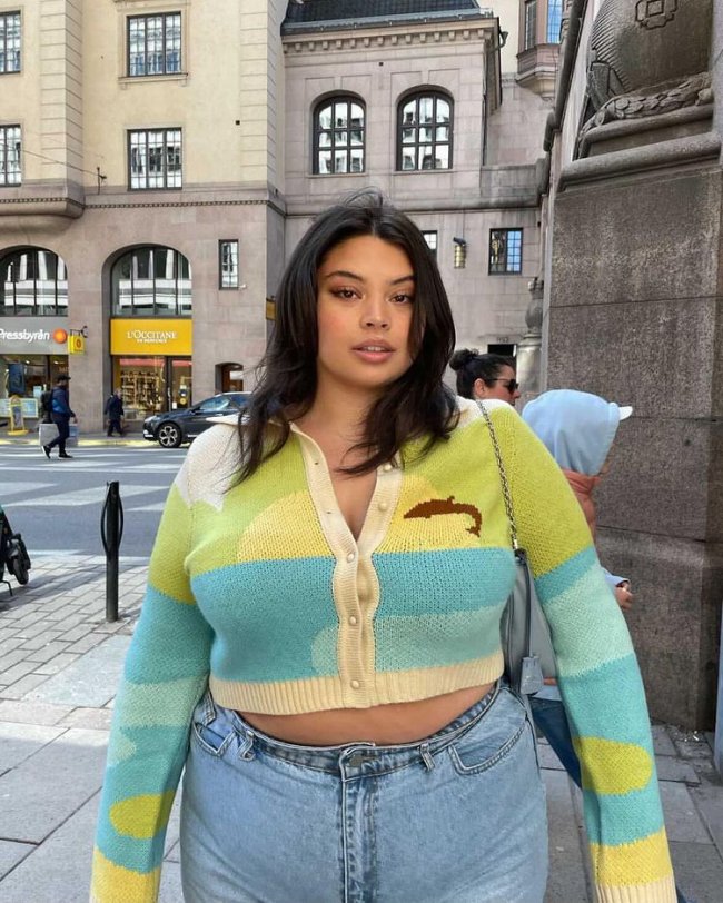 cardigan avant basic Foto de uma mulher na calçada. Ela usa um cardigan colorido em tons de verde e azul e uma calça jeans. Ela olha para a câmera e não sorri.