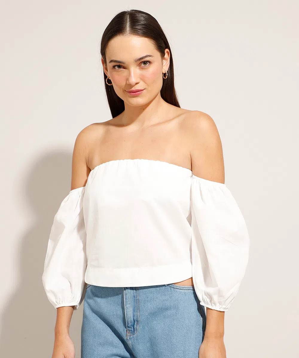 Modelo usando blusa cropped branca com mangas bufantes e, na foto, dá para ver uma parte da calça jeans. Ela está olhando para a câmera com expressão facial séria.