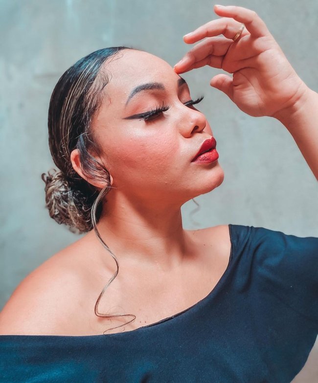 Foto de jovem com os olhos fechados, a cabeça levemente inclinada para cima, uma de suas mãos está apoiada em sua testa, sua expressão é séria. Ela usa blusa preta.