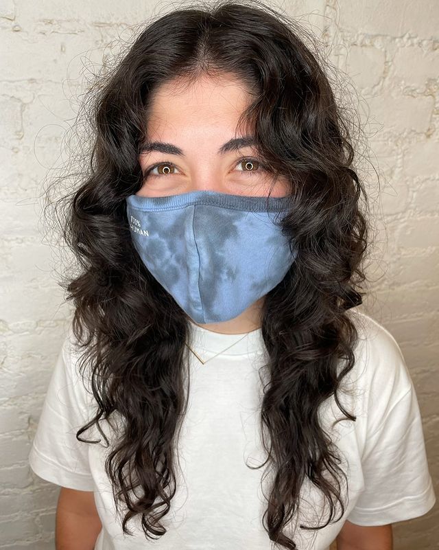 Jovem usando máscara de proteção azul e cobrindo sua expressão facial. Ela veste uma camiseta branca, pelo seu olhar parece sorridente.