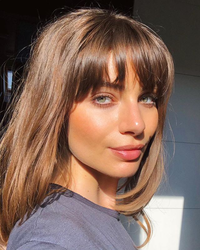 franja desfiada Selfie de uma mulher de perfil sorrindo levemente para a câmera. Ela usa uma blusa cinza, maquiagem natural e cabelo com franja desfiada solto.