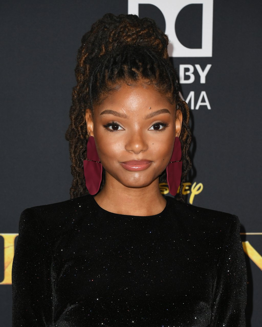 Halle Bailey na premiere de O Rei Leão; a atriz usa uma roupa preta, sorri levemente e usa brincos rosa escuro