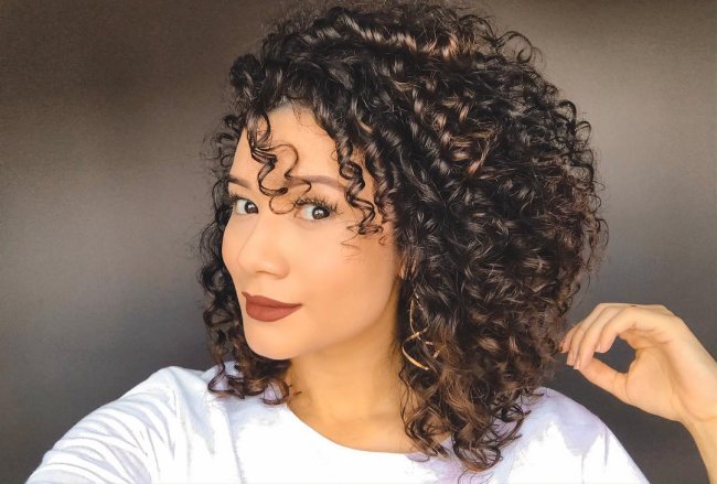 long-bob-cacheado-assimetrico Jovem com expressão levemente sorridente, exibindo seu corte bob, usando blusa branca e com a cabeça levemente inclinada