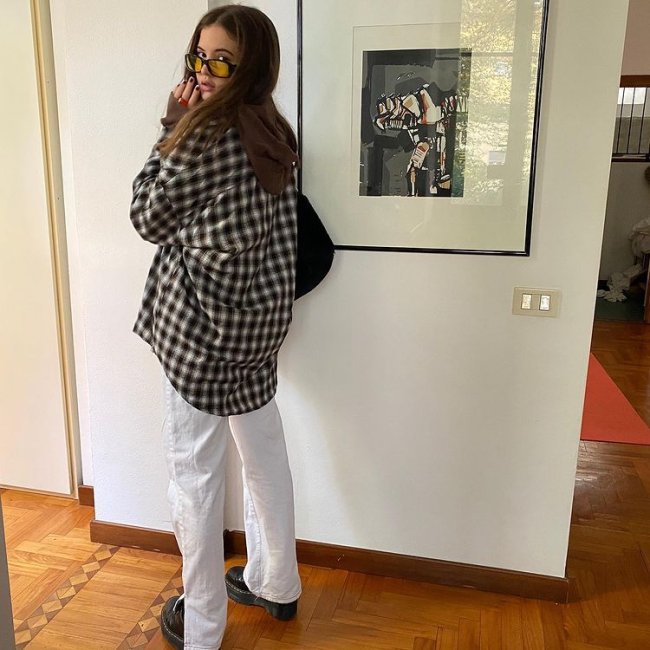 Foto de uma mulher em frente a uma parede de uma casa com um quadro. Ela usa uma camisa xadrez oversized, calça branca, coturno e óculos de sol de lente amarela. Ela está de lado, olha para a câmera e não sorri.