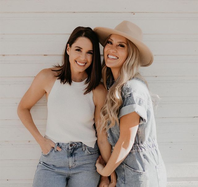 Casal posando em frente a parede branca, a menina usa blusa sem mangas branca e calça jeans clara, e a outra menina usa chapéu e macacão jeans. As duas aparecem abraçadas e sorridentes.