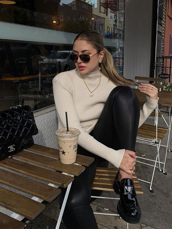 look com mocassim tratorado Jovem sentada olhando para o lado com expressão séria, ela está sentada em uma cadeira de madeira, em sua frente tem uma mesinha de madeira e ela usa blusa bege de gola alta e legging preta com mocassim preto.