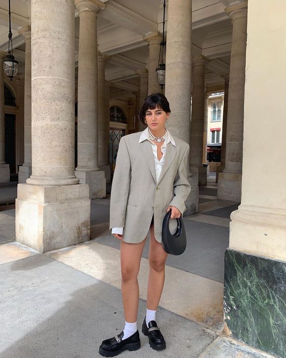 Jovem com expressão séria, posando em frente a colunas, usando blazer alongado cinza, camisa branca, e mocassim preto.