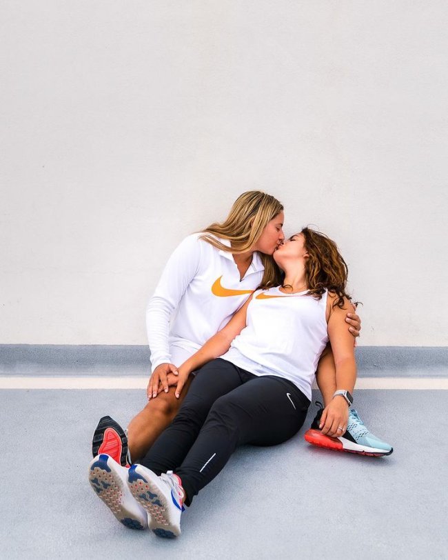 look combinando casal com camisa branca Casal deitado no chão usando roupas para academia da Nike, elas estão se beijando e não conseguimos ver seus rostos. Uma está com uma camiseta branca com manga longa da nike e a outra com regata branca da Nike e legging.