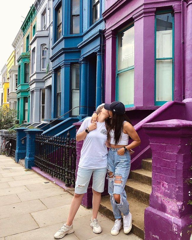 look combinando casal gay Um casal se beijando em uma rua com casas coloridas nas cores verde, cinza, azul e roxo. No casal uma moça veste camiseta branca e short, e a outra boné preto, cropped branco e calça destroyed. Não conseguimos ver o rosto delas.