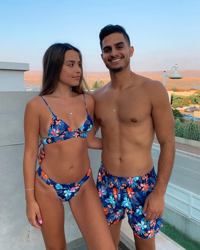 look praia Casal abraçado com trajes de banho, a menina usa biquini azul com detalhes coloridos, e o menino usa bermuda com a mesma estampa do biquini dela. Ela aparece com o rosto virado para o lado direito e ele olha sorridente para frente.