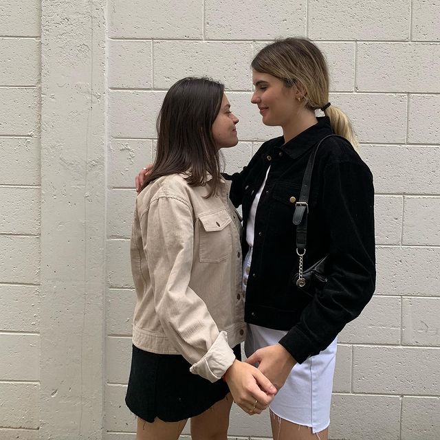 looks combinando Casal posando em frente a parede branca de tijolos, se olhando. As duas aparecem de perfil. Uma usa jaqueta bege com saia preta e a outra sobreposição de blusa preta e saia branca.