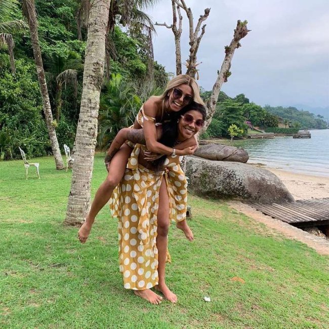 ludmilla Ludmilla e Bruna posando em um local com grama e arvores. Bruna está montada nas costas de Ludmilla e as duas aparecem sorridentes.