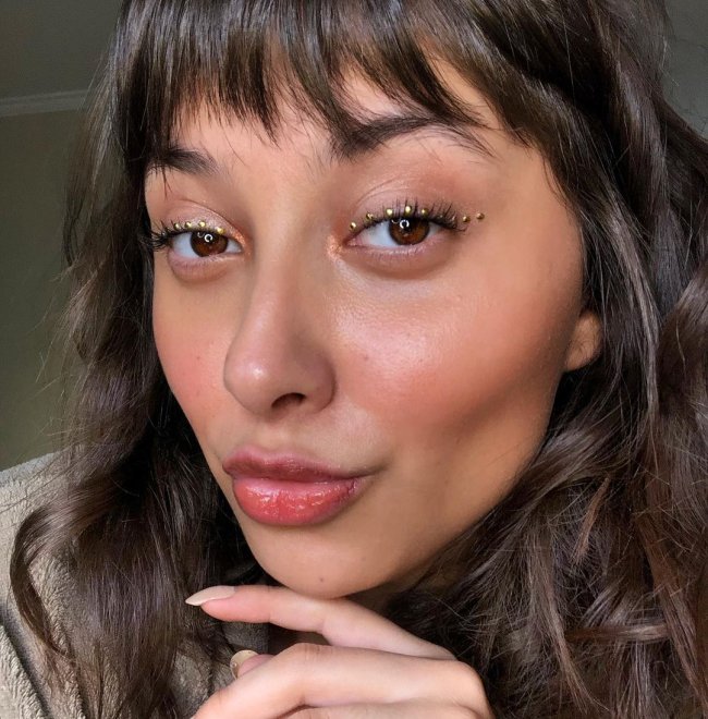 make com strass Jovem fazendo biquinho para selfie, com expressão levemente sorridente, usando strass nos olhos e pele com pouca maquiagem.