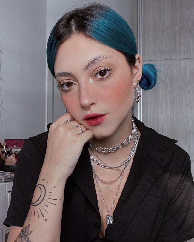 maquiagem cantinho do olho iluminado Jovem tirando selfie com a cabeça levemente inclinada, ela tem cabelo azul preso em coque e sua expressão é séria. Ela usa colares pratas e uma blusa preta.