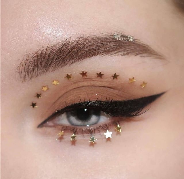 maquiagem com estrelinhas Foto com foco em um olho aberto, com delineado preto e estrelinhas douradas ao redor dele.