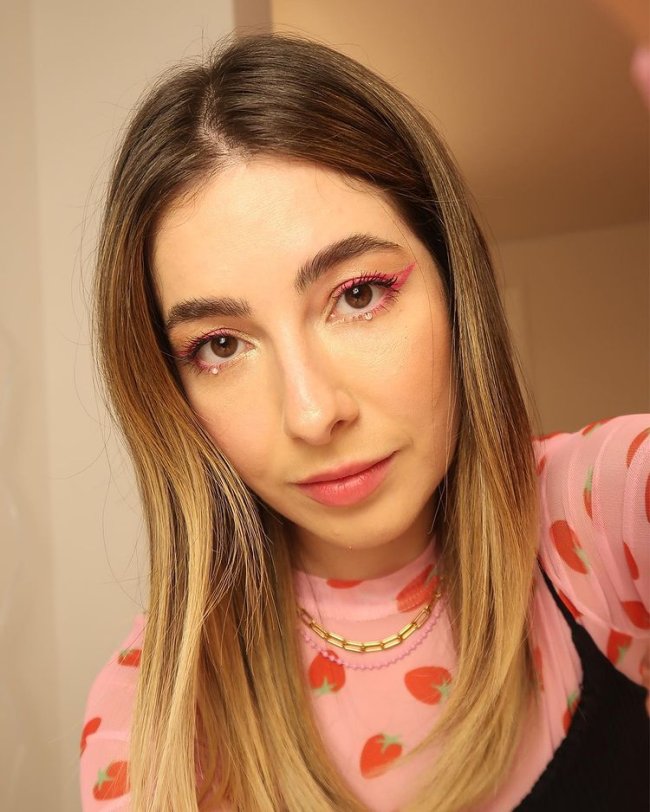 maquiagem com strass Jovem tirando selfie usando blusa segunda pele rosa com corações vermelhos, regata preta, delineado colorido e com expressão séria.