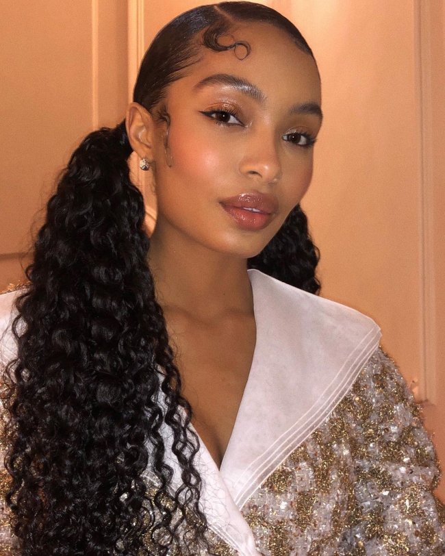 maria chiquinha cabelo cacheado Foto de uma mulher com o penteado maria chiquinha baixo. Ela usa um casaco com detalhes dourados e pérolados, está com uma make glow e brinco simples. Ela olha para a câmera e sorri levemente.