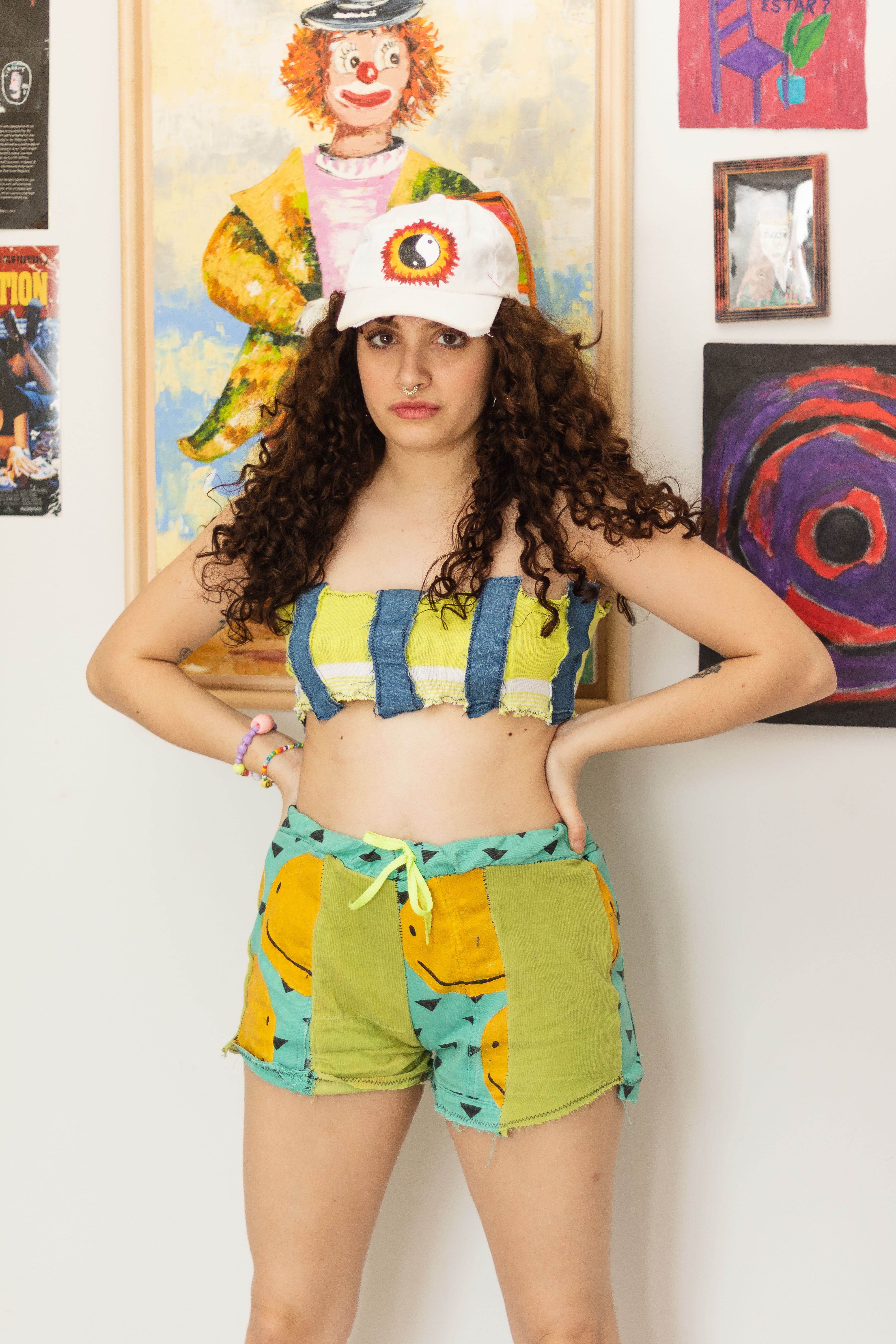 melissa-achei-super-cool-marca-sustentavel-upcycling Melissa Rodrigues, criadora da marca Achei Super Cool. Ela está em pé, usando um top cropped jeans e amarelo, um short colorido com carinhas smile e um boné branco com símbolo yin-yang. Ela está com expressão facial séria e as duas mãos na cintura.