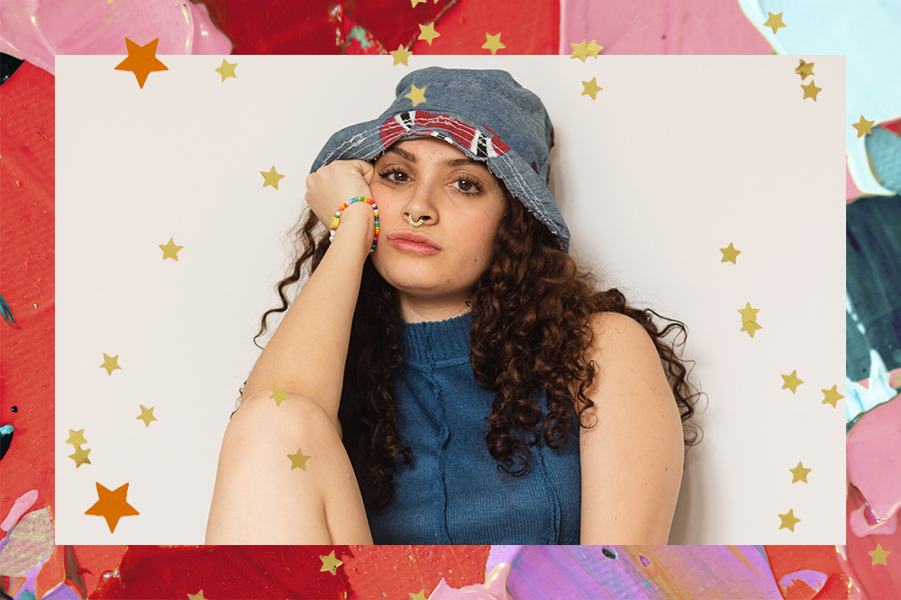 Melissa Rodrigues, criadora da marca Achei Super Cool. Ela está usando um bucket hat jeans e um top cropped jeans. Está sentada com uma das pernas dobradas e um dos braços apoiados no rosto. A montagem tem fundo com tintas coloridas e estrelinhas.