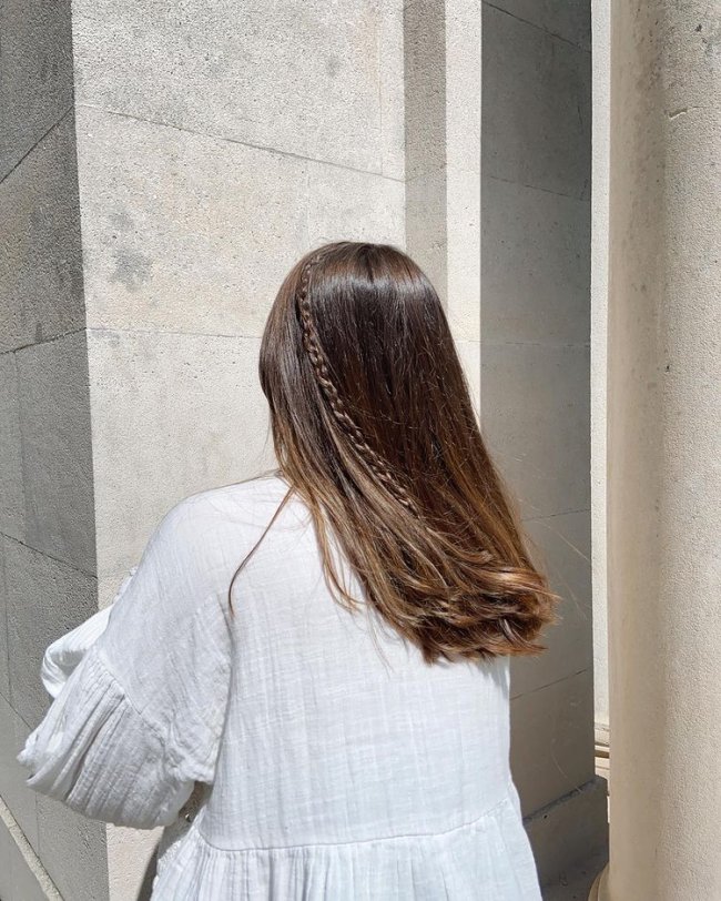 mini trança Foto de uma mulher de costas com uma mini trança no cabelo. Ela usa uma blusa branca e está com o cabelo liso solto.