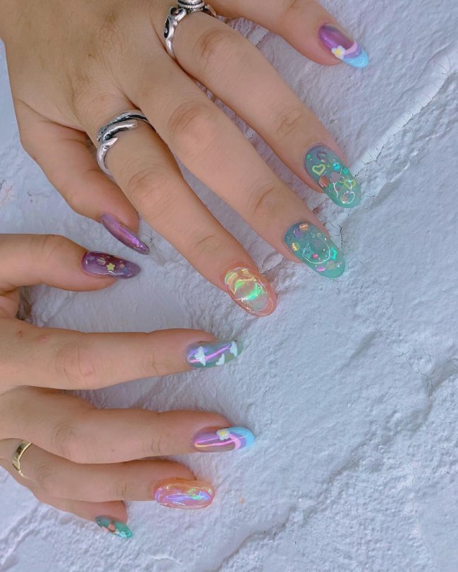 Foto com foco nas unhas, duas mãos expostas em mesa branca. Com nail art nas cores azul, amarelo, verde, e rosa.