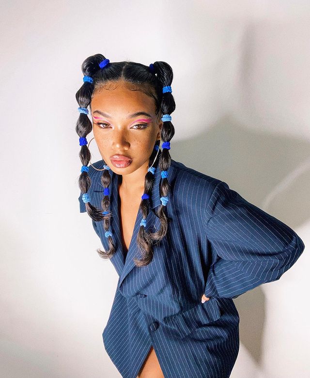 penteado bolha colorido Foto de uma mulher em frente a uma parede branca. Ela usa um blazer azul com riscos brancos, cabelo preso em um penteado bolha dividido em quatro partes. Ela está com as mãos na cintura, olha para a câmera e não sorri.