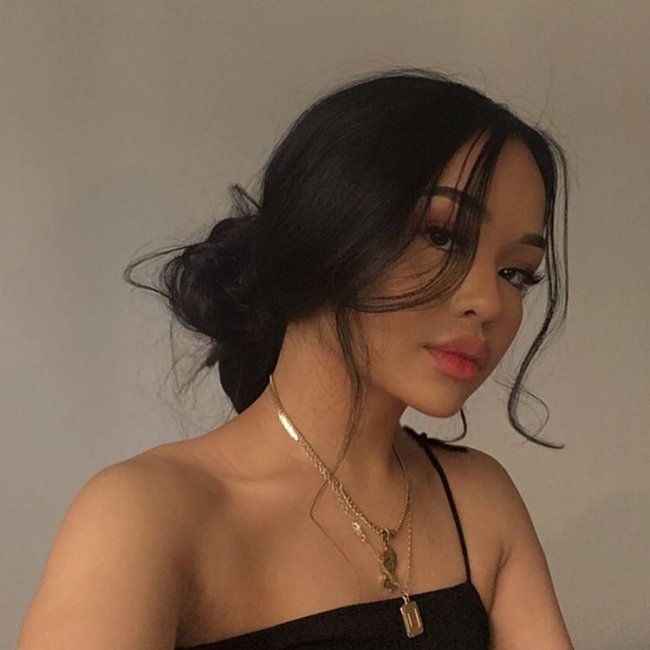 penteado coque baixo Selfie de uma mulher. Ela usa um cropped preto, colares dourados e o cabelo em um coque baixo bagunçadinho. Ela olha para a câmera e não sorri.