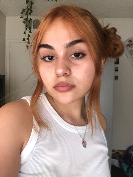 penteado space buns Selfie de uma garota. Ela usa uma blusa branca, colar dourado e cabelo preso em dois coques laterais, o penteado space buns. Ela olha para a câmera e não sorri.