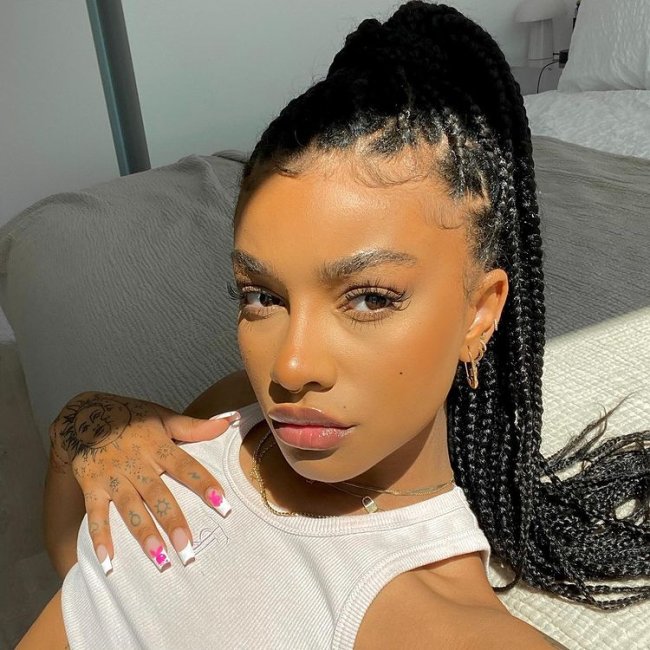 rabo de cavalo com trança Selfie de uma mulher com o penteado rabo de cavelo no cabelo trançado. Ela usa um top cropped branco, brinco e colares dourados. Ela está com a mão direita apoiada no seio, está deitada no sofá, olha para a câmera e não sorri.