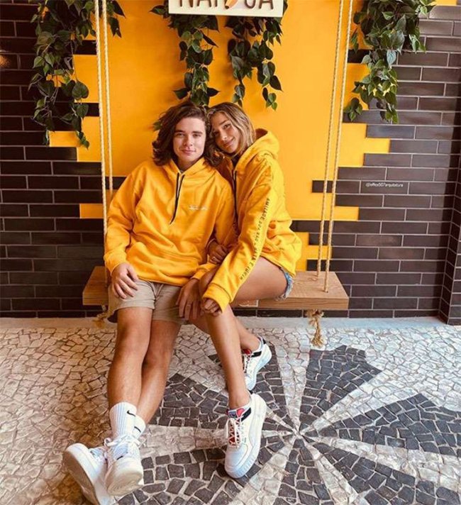 Sasha e Marido Casal sentado em balanço usando roupa combinando. Blusa de moletom amarela e short com tênis branco. Os dois aparecem sorridentes.