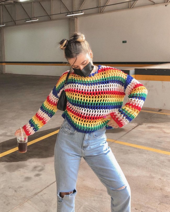 space buns lorrine mondin Foto da influencer Lorrine Mondin em um estacionamento. Ela usa uma blusa de tricô colorida, calça jeans, máscara preta e penteado space buns. Ela está segurando um copo com a mão direita, com a mão esquerda na cintura e olha para baixo.