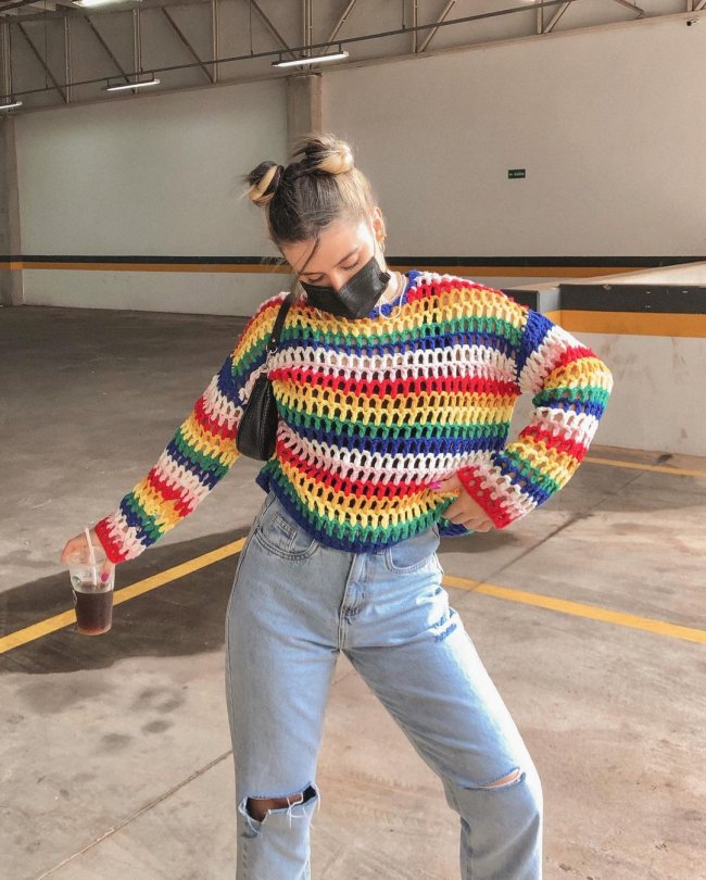 Foto da influencer Lorrine Mondin em um estacionamento. Ela usa uma blusa de tricô colorida, calça jeans, máscara preta e penteado space buns. Ela está segurando um copo com a mão direita, com a mão esquerda na cintura e olha para baixo.