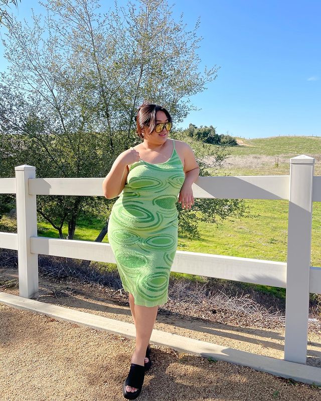 vestido avant basic Foto de uma mulher em frente a uma cerca. Ela usa um vestido verde menta com estampa oval em tons de um verde mais escuro, óculos de sol com lente amarela, tamanco preto e cabelo preso em um coque. Ela olha para o lado e sorri levemente.