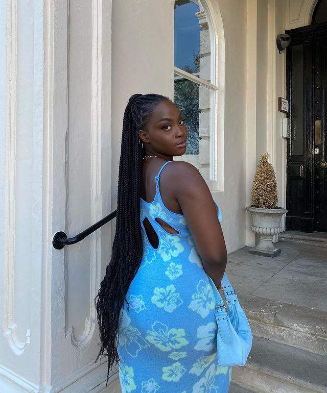 vestido azul avant basic Foto de uma mulher em frente a uma casa. Ela usa um vestido azul com estampa de flores em um tom de azul mais claro e segura uma bolsa azul. Ela olha para a câmera e não sorri.