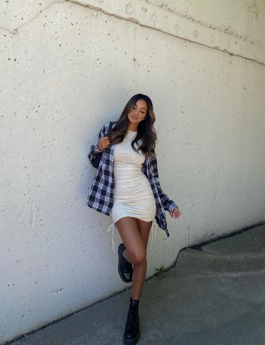 vestido e camisa xadrez Foto de uma mulher na rua. Ela usa um vestido na cor creme, camisa xadrez e coturno. Ela está com a perna direita levemente levantada, olha para baixo e não sorri para a foto.