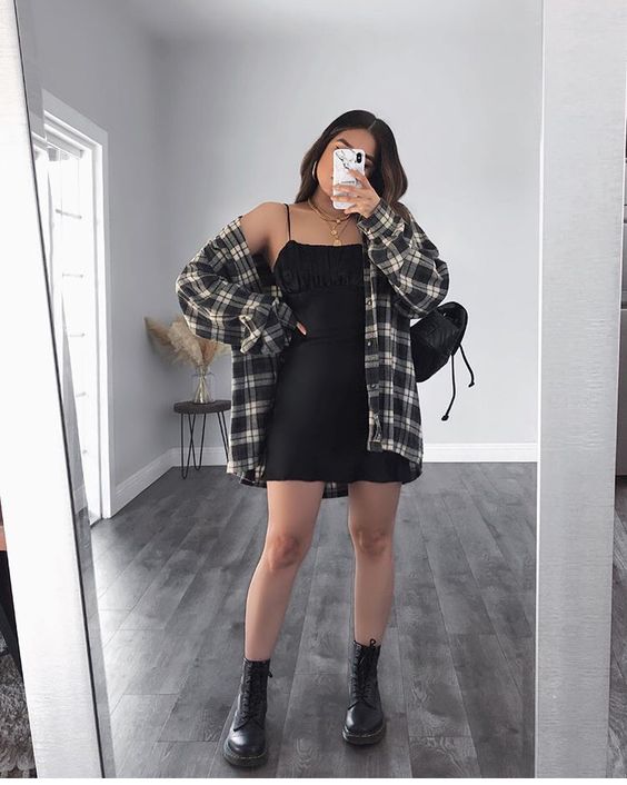 vestido preto e camisa xadrez Selfie no espelho de uma mulher. Ela usa um vestido preto curto, camisa xadrez oversized, coturno preto e bolsa preta.