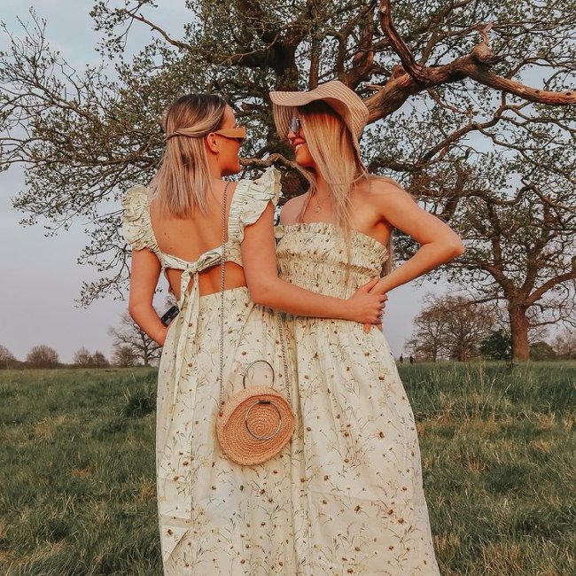 vestidos combinando casal Casal posa em frente a arvores em chão com gramado, as duas estão se olhando e o cabelo cobre suas expressões faciais. Uma está de lado para a câmera e a outra de costas. As duas usam vestido com estampa floral igual.