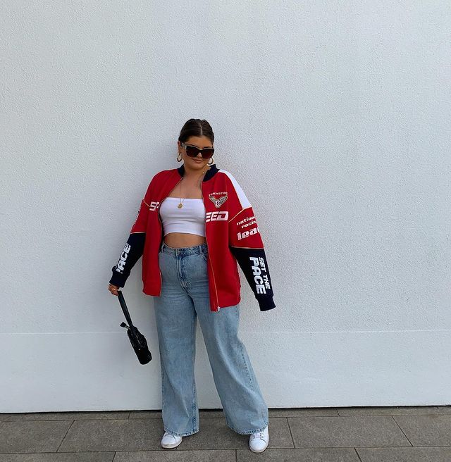 Garota usando top cropped branco, calça jeans wide leg, tênis branco e jaqueta oversized vermelha, preta e branca. Ela está segurando uma bolsa preta com uma das mãos, olhando para baixo e usando um óculos de sol.