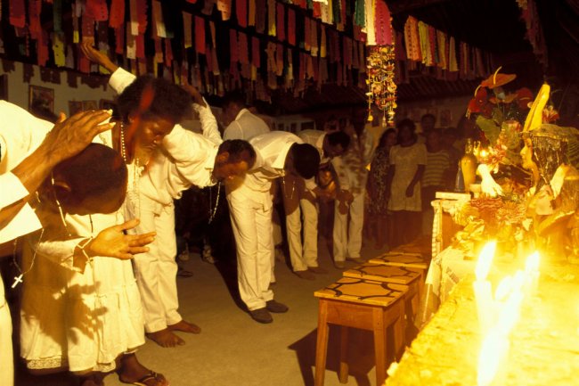 A Intolerância religiosa, o racismo e a síndrome da superioridade europeia Imagem mostra um culto de candomblé realizado na Bahia. Negros vestidos de brancos se curvam em frente a estátuas de santos.