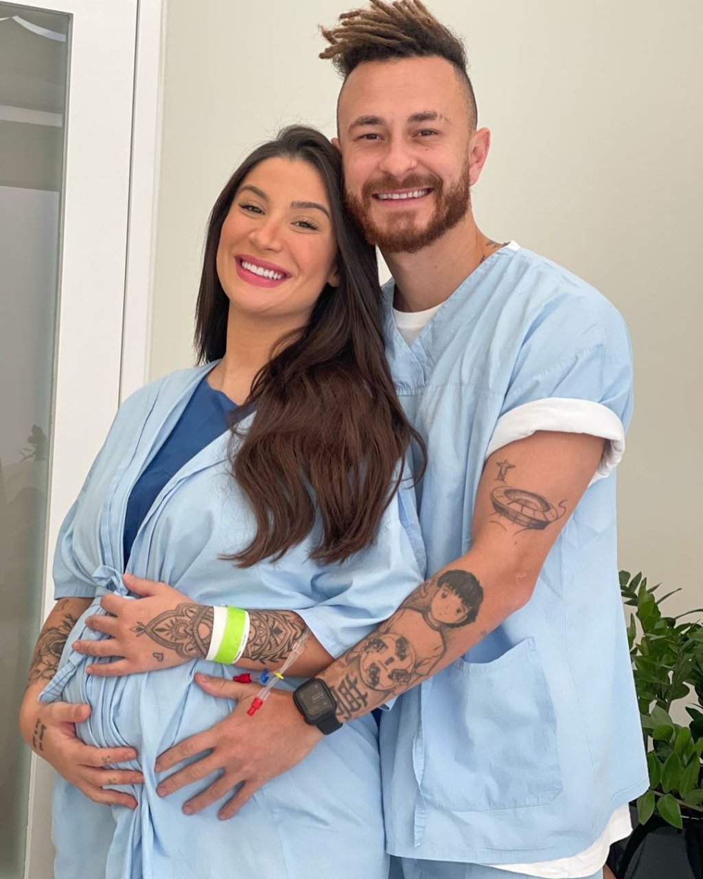 Bianca Andrade e Fred na maternidade; os dois estão usando roupas azuis de hospital e Fred está abraçando Bia por trás com as mãos na barriguinha de grávida dela; Bia também está com as mãos na barriga